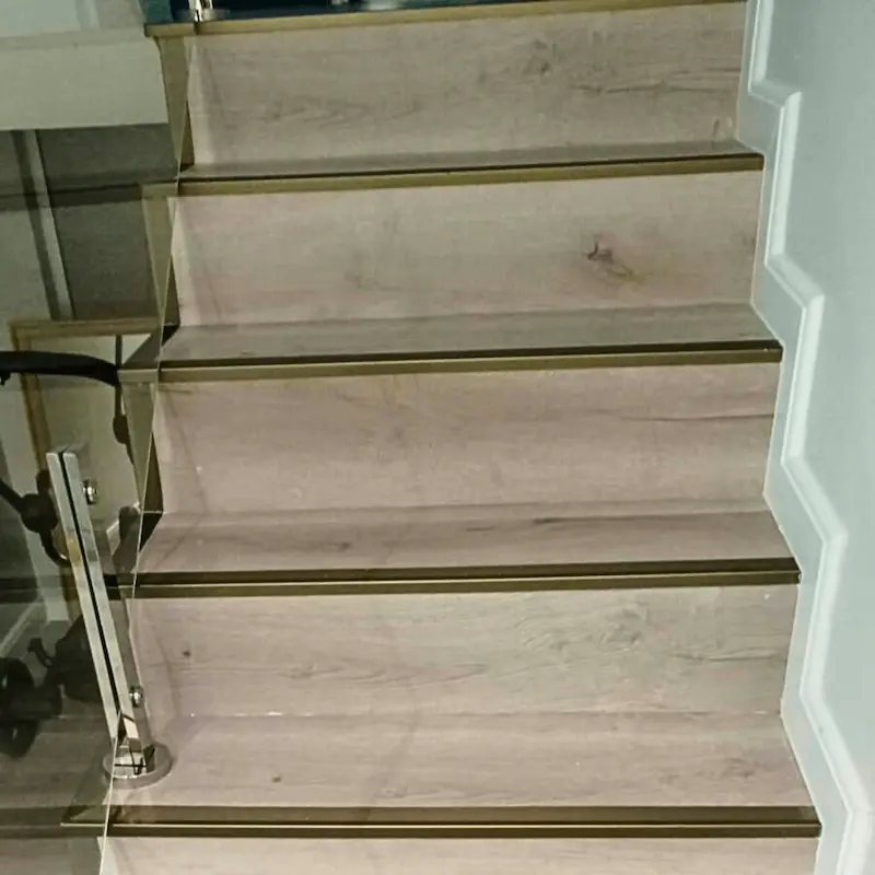 Detalhe dos degraus da escada com revestimento amadeirado e perfil de acabamento metálico antiderrapante.